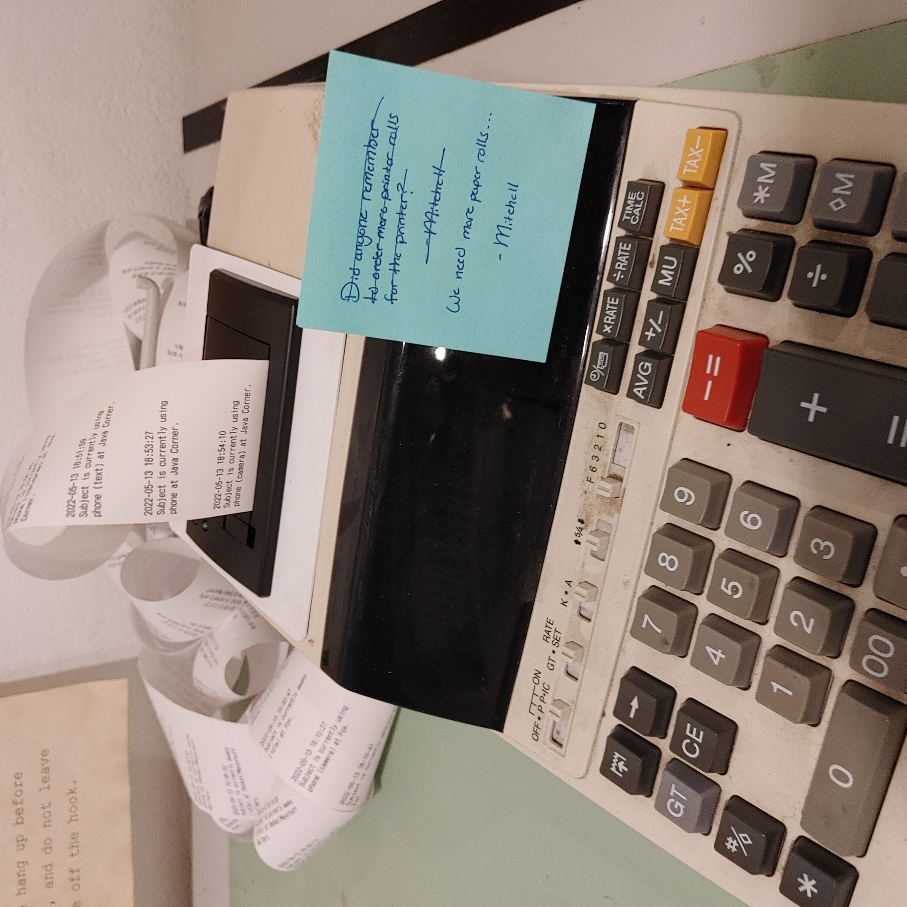 Photo of the accounting calculator close up. The logged activities of PT are legible on the spooled out paper. A blue post-it note asking for more paper is stuck to the front of the calculator.